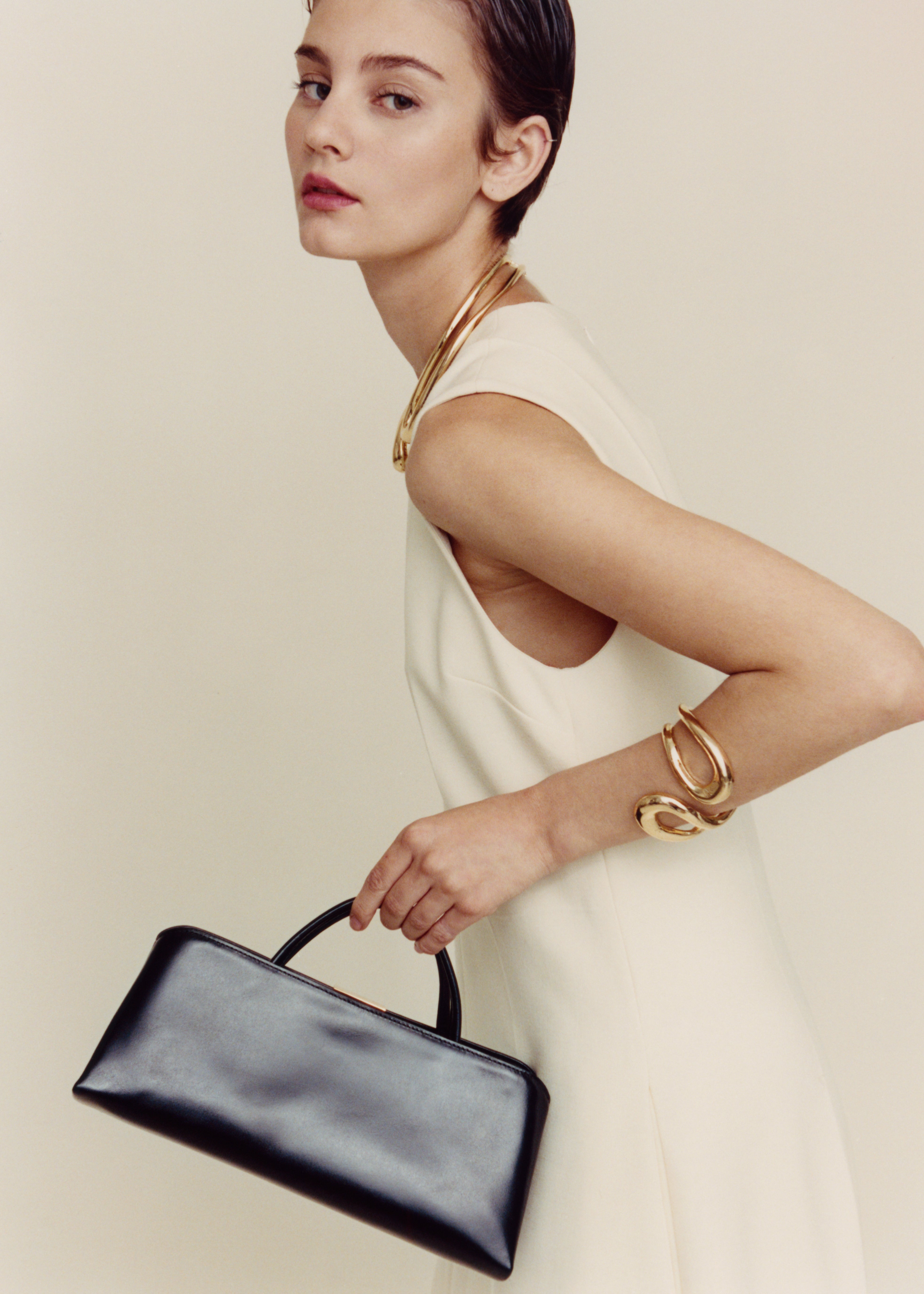 Woman holding a Gold Noir Éclair Clutch Shoulder Handbag (Black) with gold jewelry against a beige background