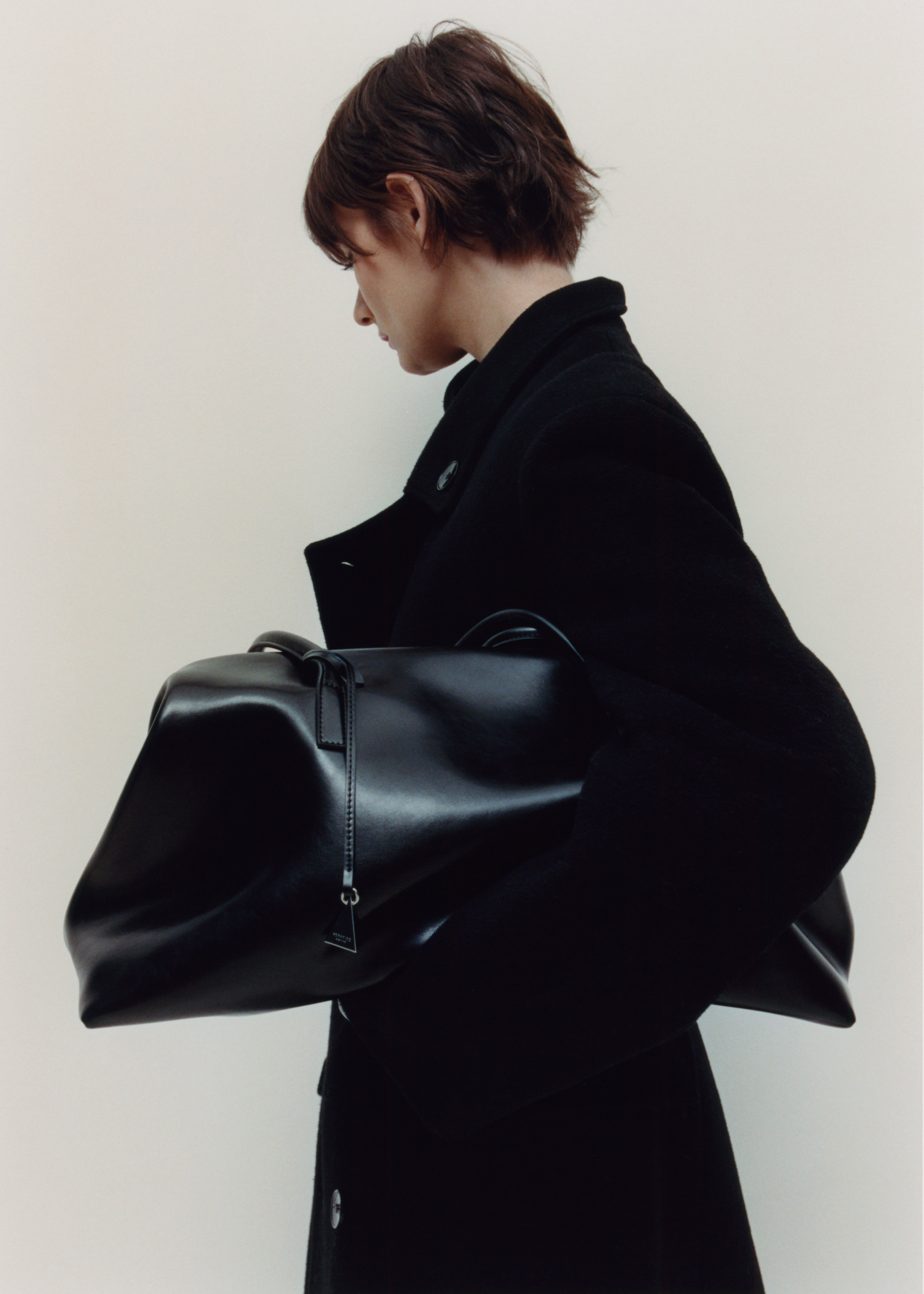 Person wearing a black coat holding a large Black Club Large Handbag against a plain background