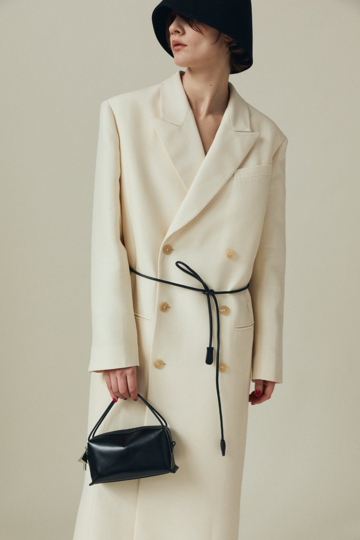 Person wearing a long cream coat with a black belt, holding a Verafied New York Black Lizard Café Mini Handbag against a neutral background.