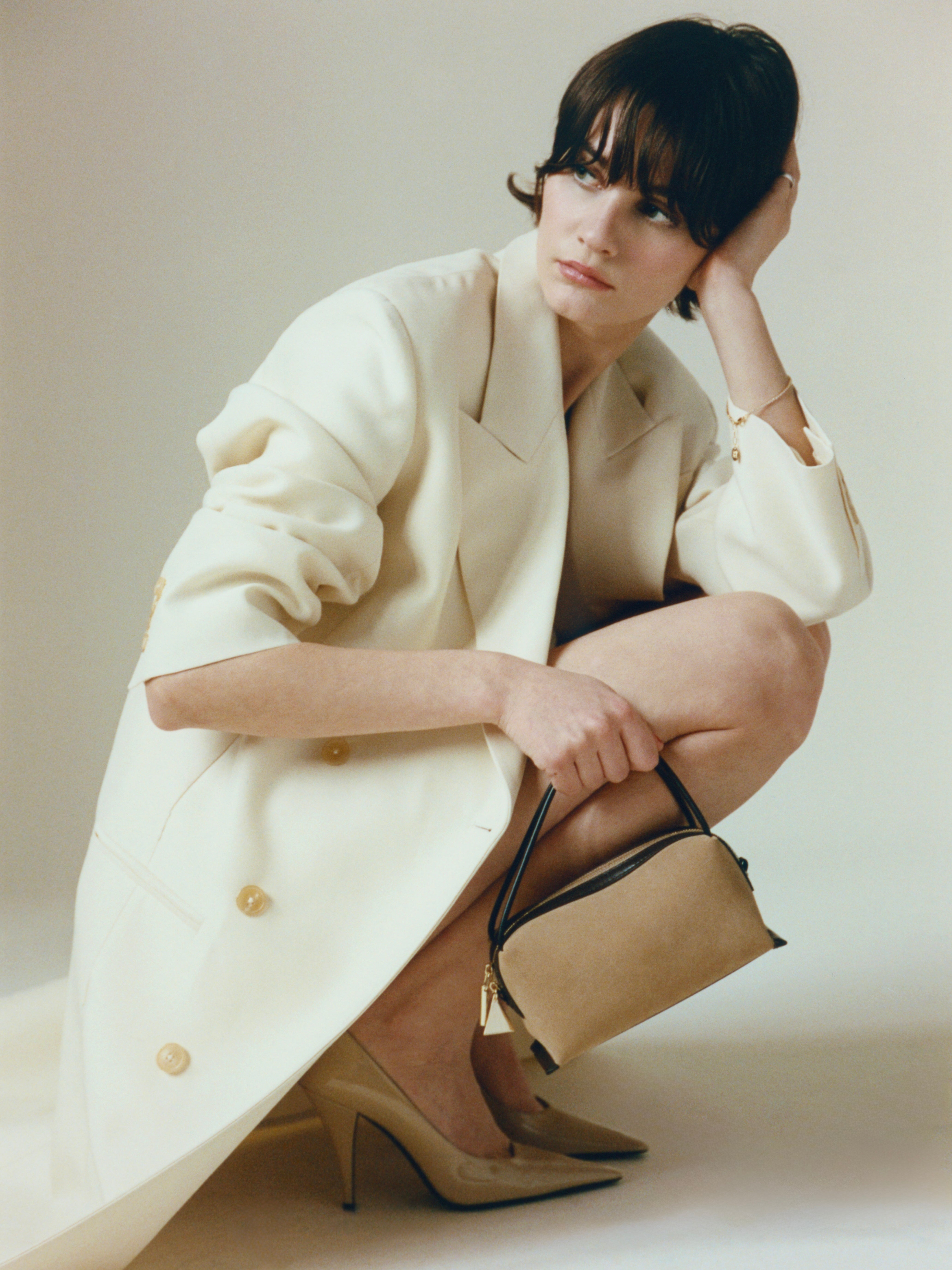 Woman in a long white coat holding a Verafied New York Gold Suede Café Mini Handbag against a plain background