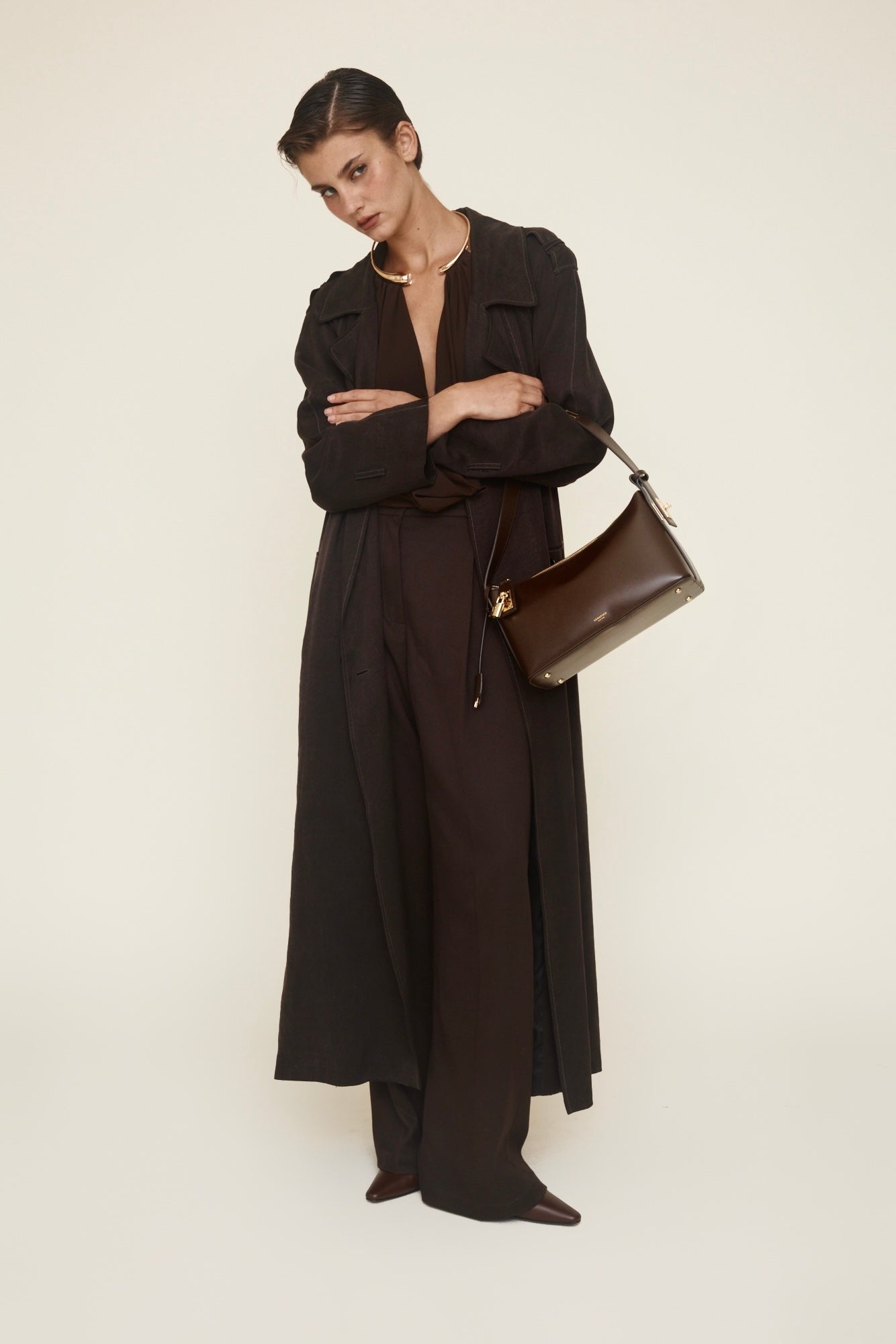 Woman in a long black coat holding a brown hobo bag against a white background