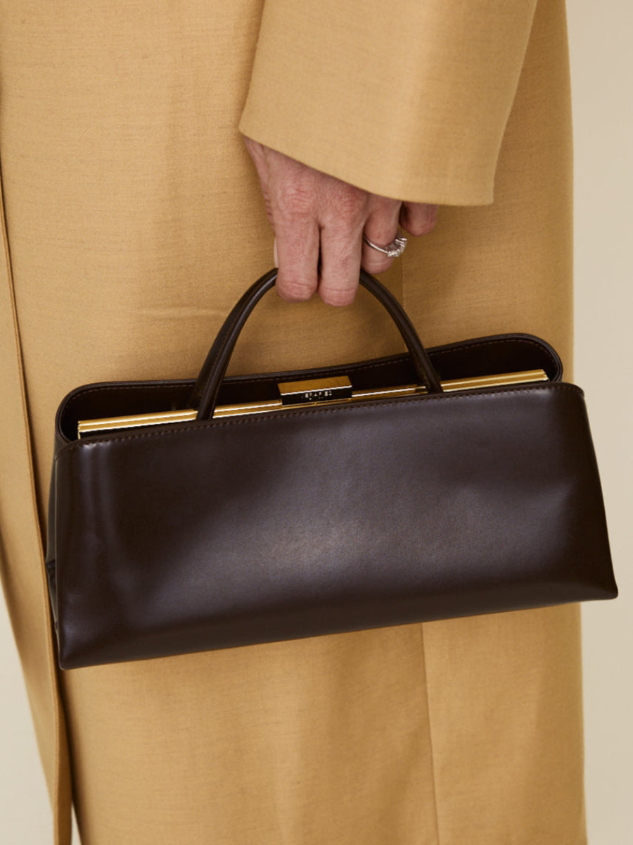 Person holding a Chocolate Éclair Clutch Shoulder Handbag with gold accents, wearing a beige coat.