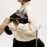 Person holding a Chocolate Medium Club Shoulder Handbag, sitting on a chair against a white background