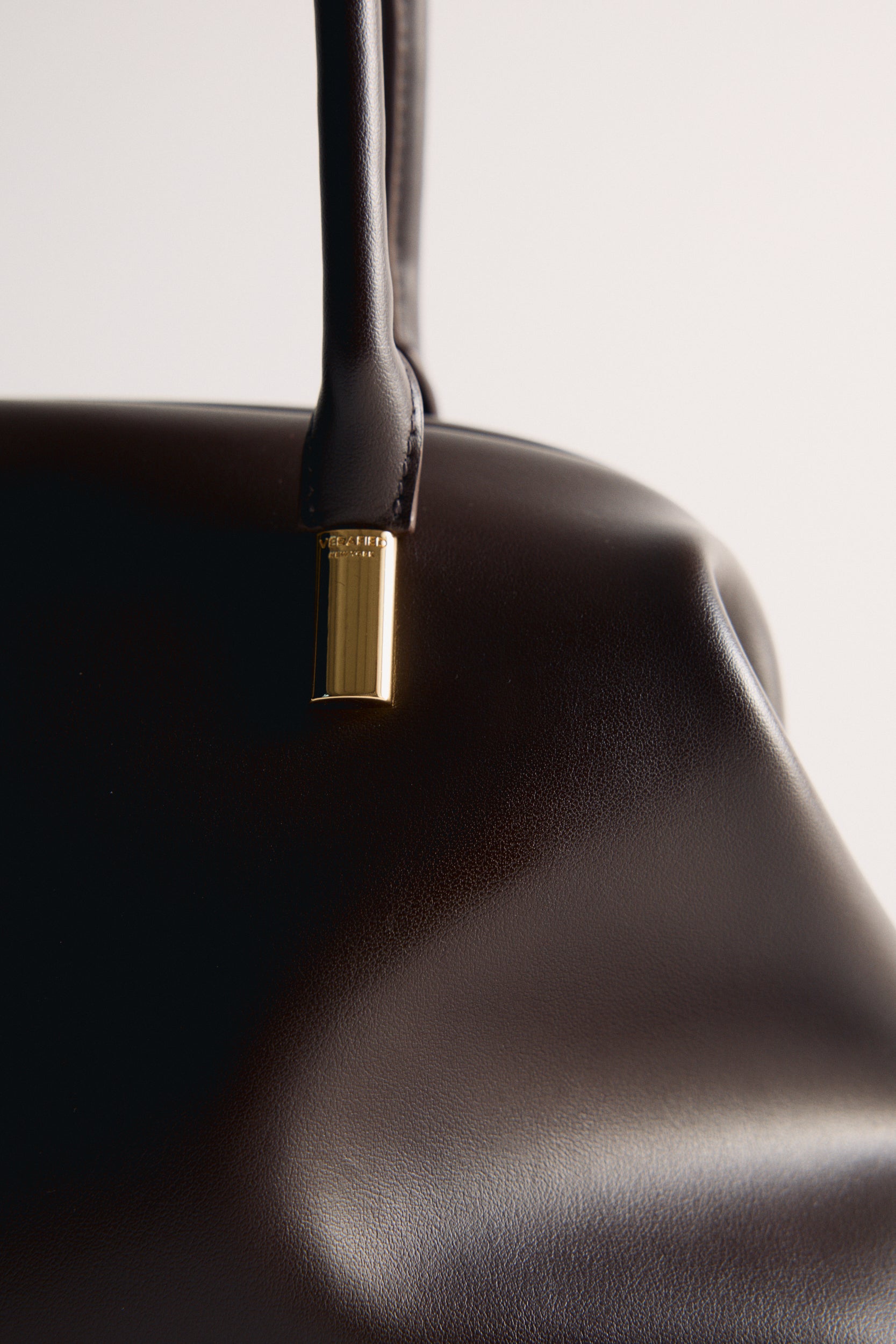 Close-up of a Chocolate Medium Club Shoulder Handbag with a gold clasp on a light gray background