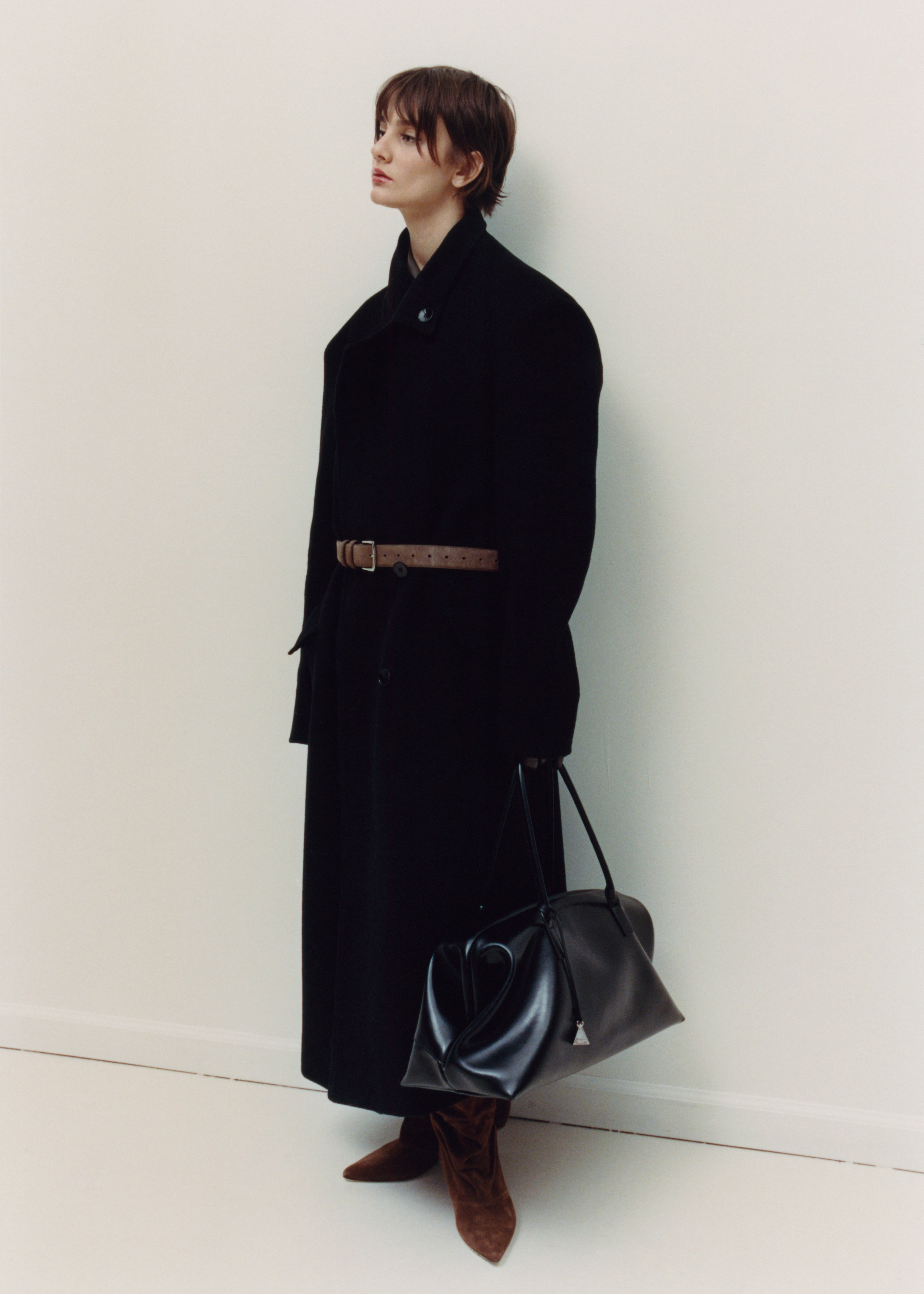 Person wearing a long black coat holding a Black Club Large Handbag against a white wall