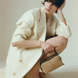 Woman in a long white coat holding a Verafied New York Gold Suede Café Mini Handbag against a plain background