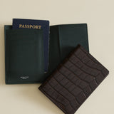 Black passport holder with a passport and Mocha Croc Embossed Passport Cover Wallet on a beige background