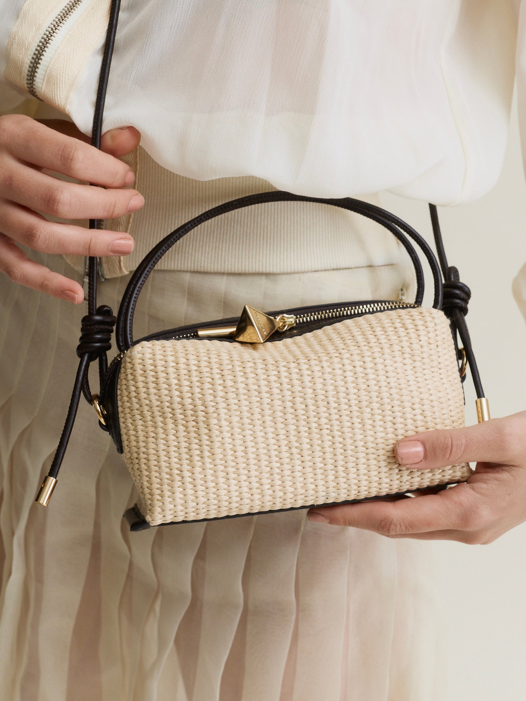 Beige Raffia Café Mini Crossbody Handbag with black straps held by a person wearing a white dress.