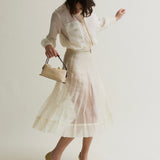 Woman wearing a light-colored outfit with a skirt and holding a Raffia Café Mini Crossbody Handbag on a plain background