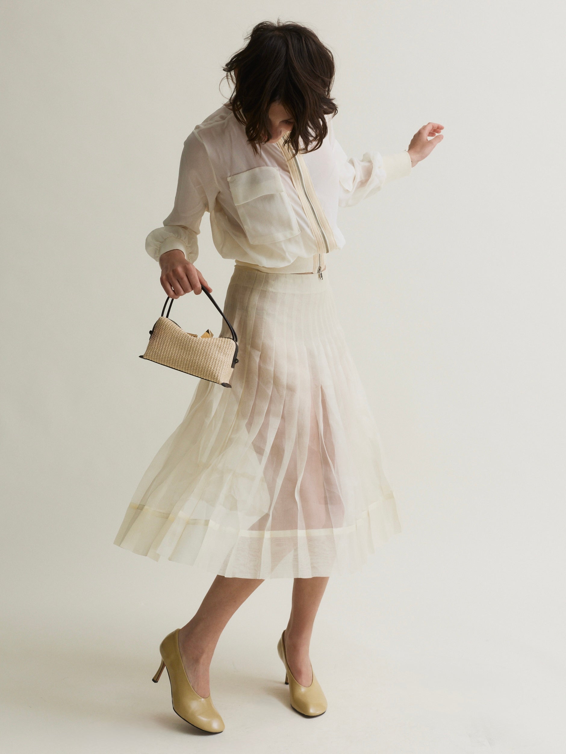 Woman wearing a light-colored outfit with a skirt and holding a Raffia Café Mini Crossbody Handbag on a plain background