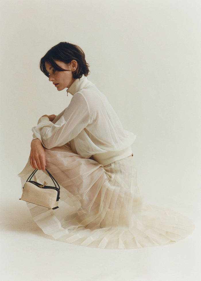 Woman in a white dress holding a Raffia Café Mini Crossbody Handbag against a plain background