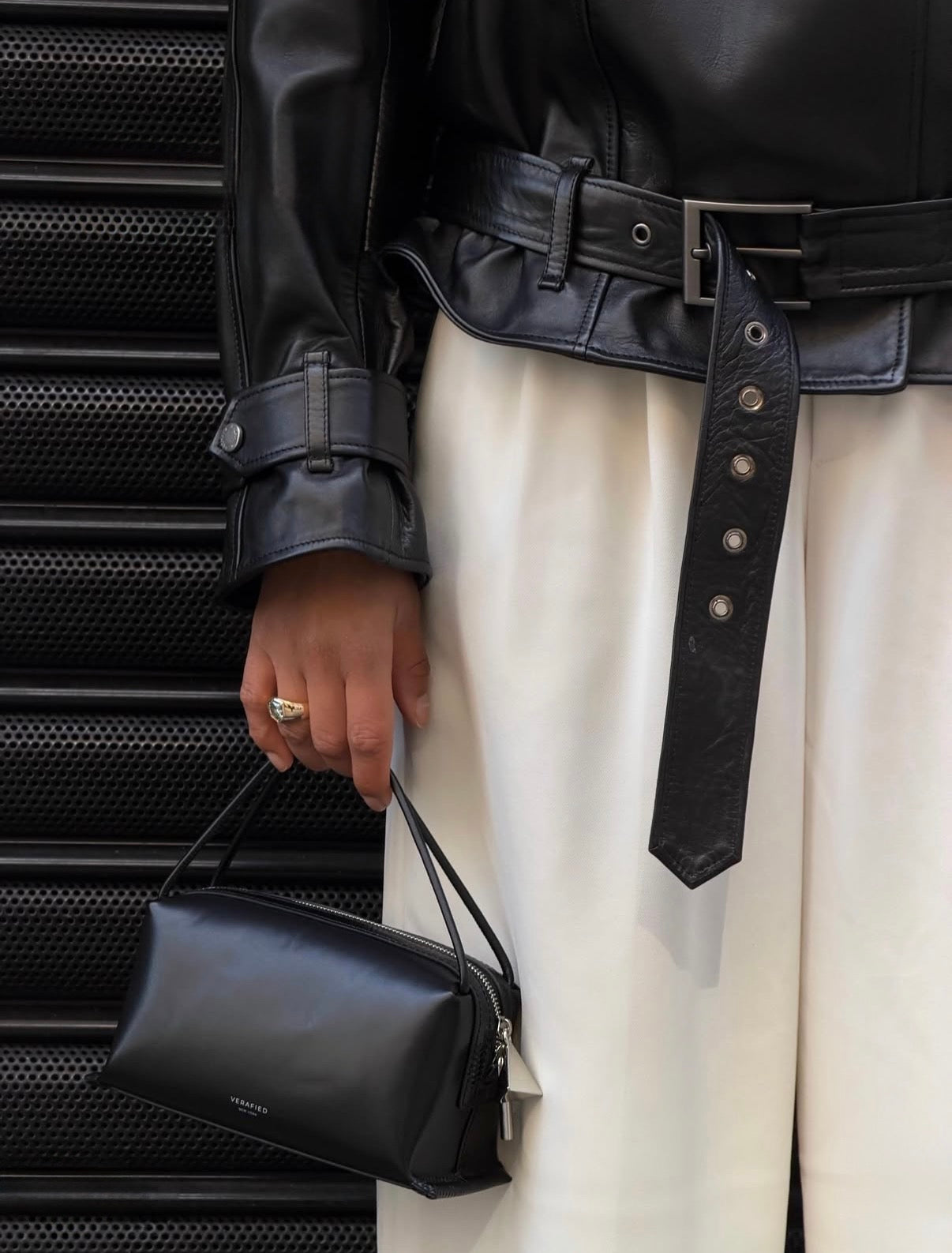 A close-up of a person wearing a black leather jacket with a wide belt and white wide-leg pants, holding a small Black Lizard Café Mini Crossbody Handbag against a dark textured metal background.