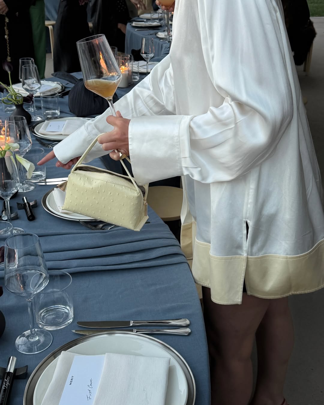 Person in a white outfit with beige trim holding a beige Butter Ostrich Café Mini Bag: Crossbody Handbag at a formal event table.
