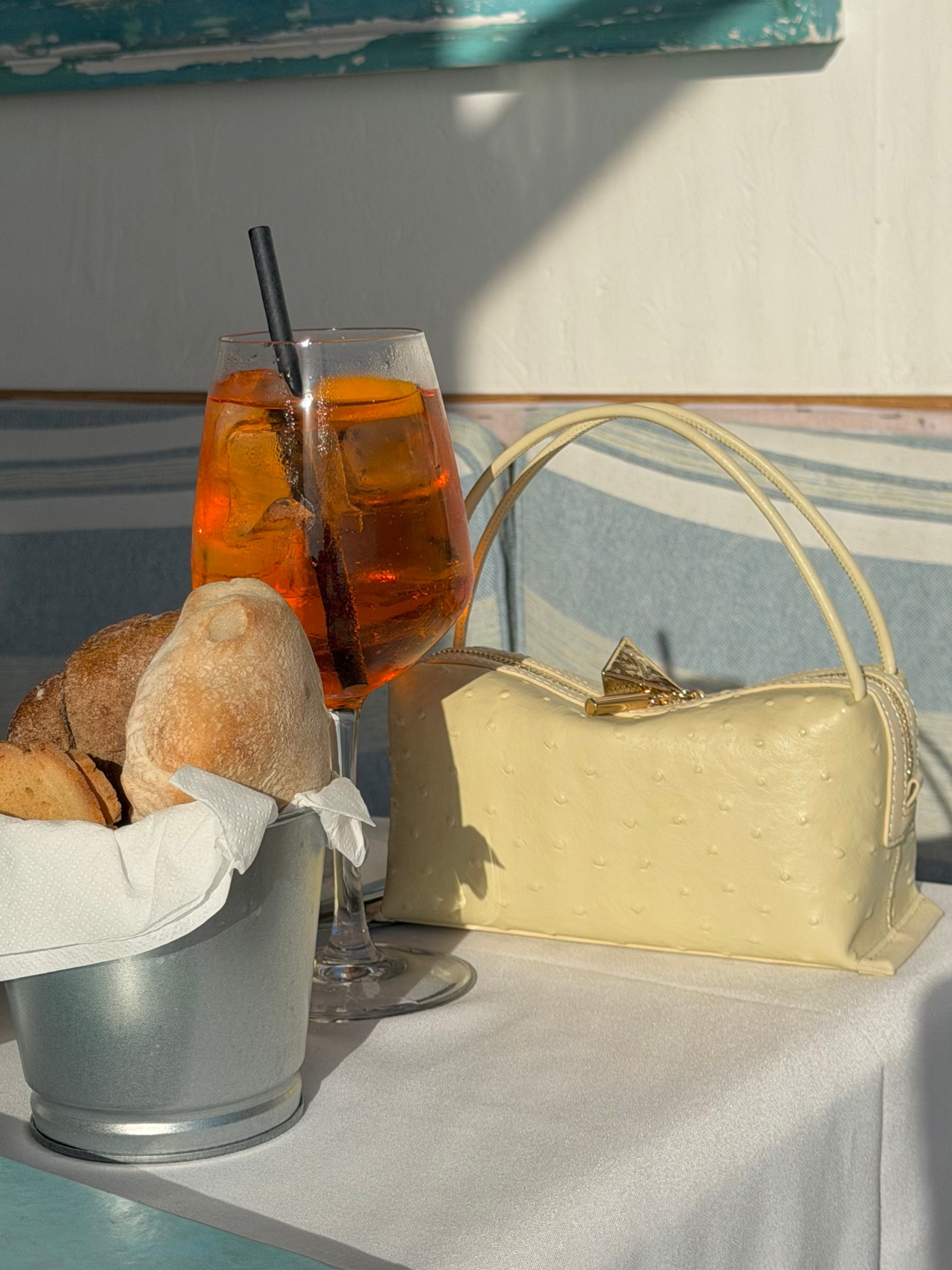 Glass of orange beverage with a straw, bread loaf, and beige Butter Ostrich Café Mini Bag: Crossbody Handbag on a table with a striped tablecloth.