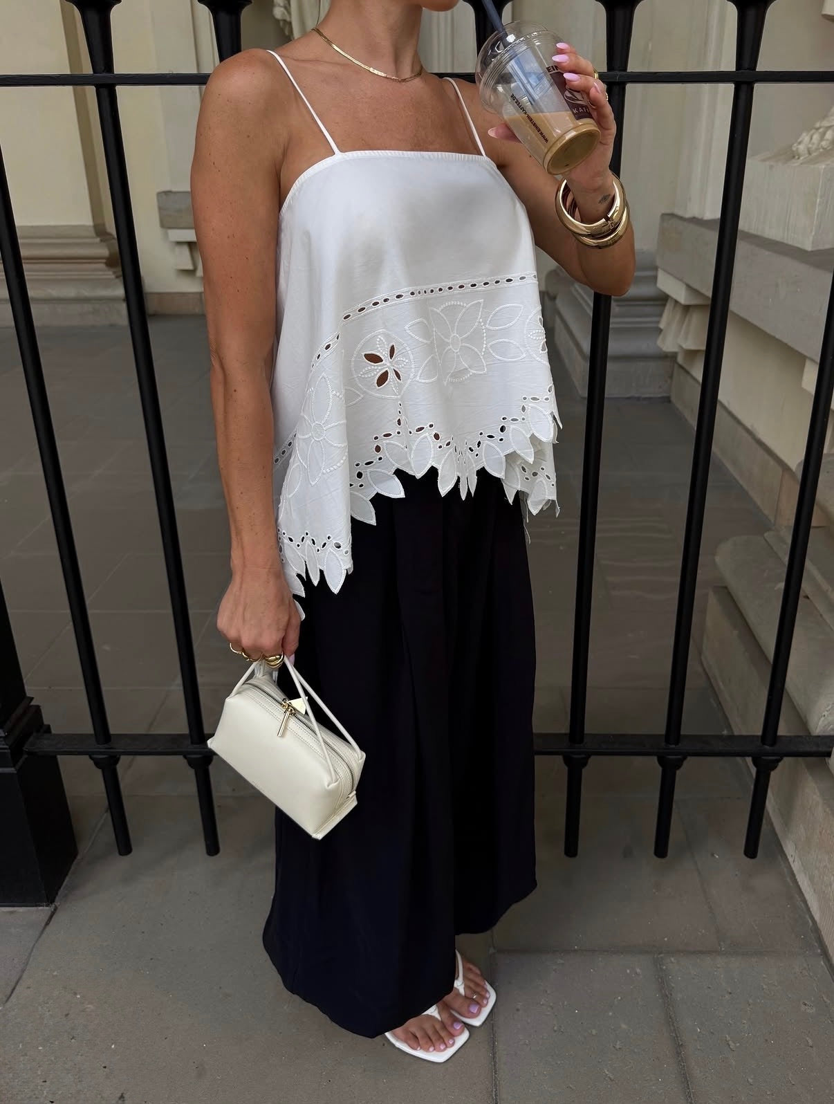A woman wearing a white spaghetti-strap top with a scalloped eyelet lace hem and a long navy skirt, holding a small Ivory Café Mini Crossbody Handbag.