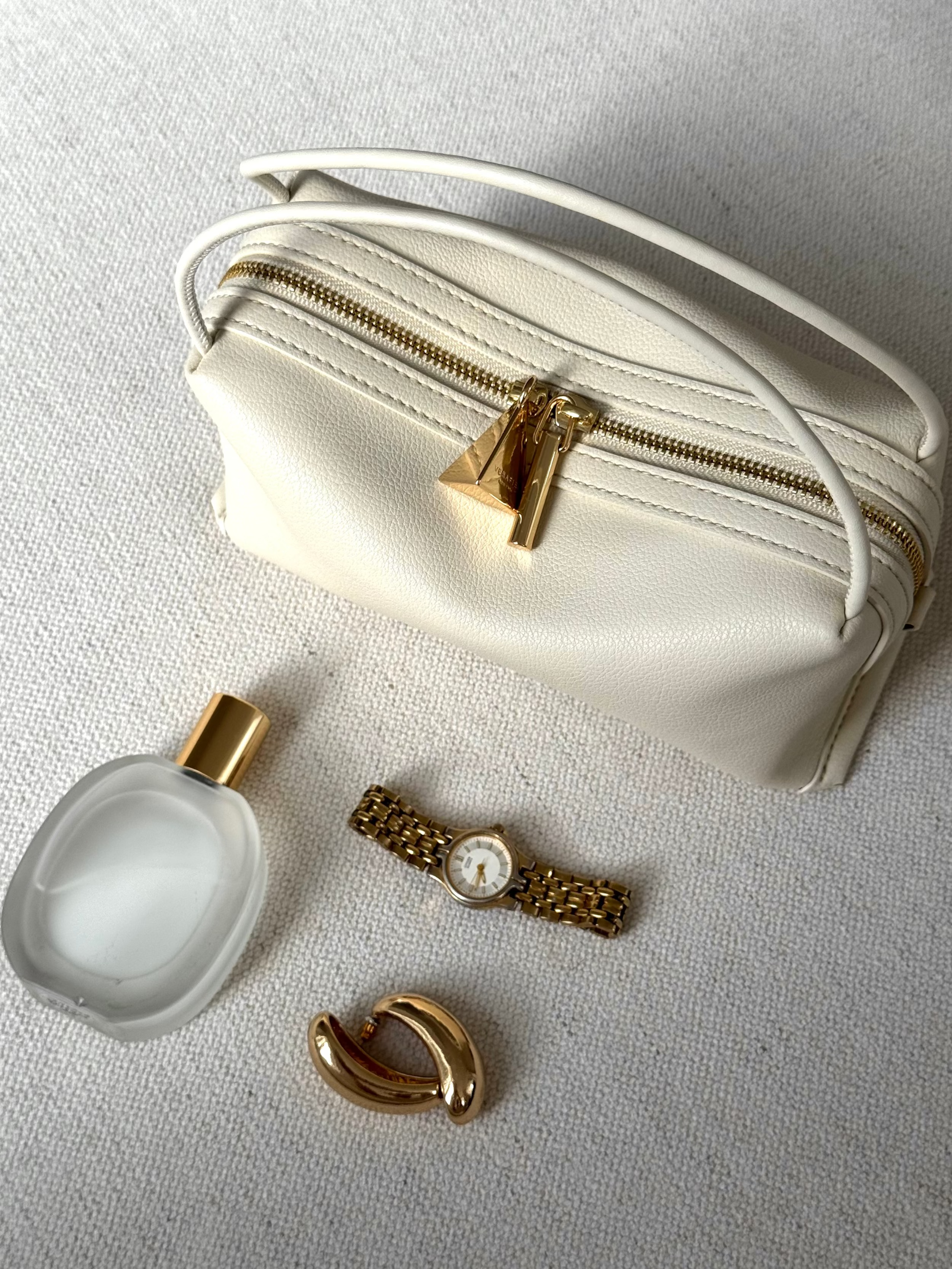 White Ivory Café Mini Crossbody Handbag with gold accents, a small white bottle, a watch, and a gold earring on a light gray background.
