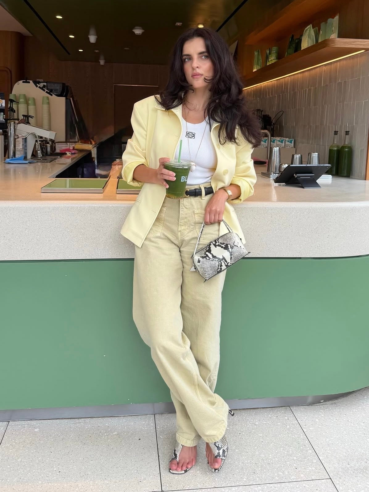 A woman stands in a cafe setting wearing a light yellow blazer, matching cream trousers, a white top, and holding a green drink. She is holding a small Python Café Mini Bag: Crossbody Handbag.