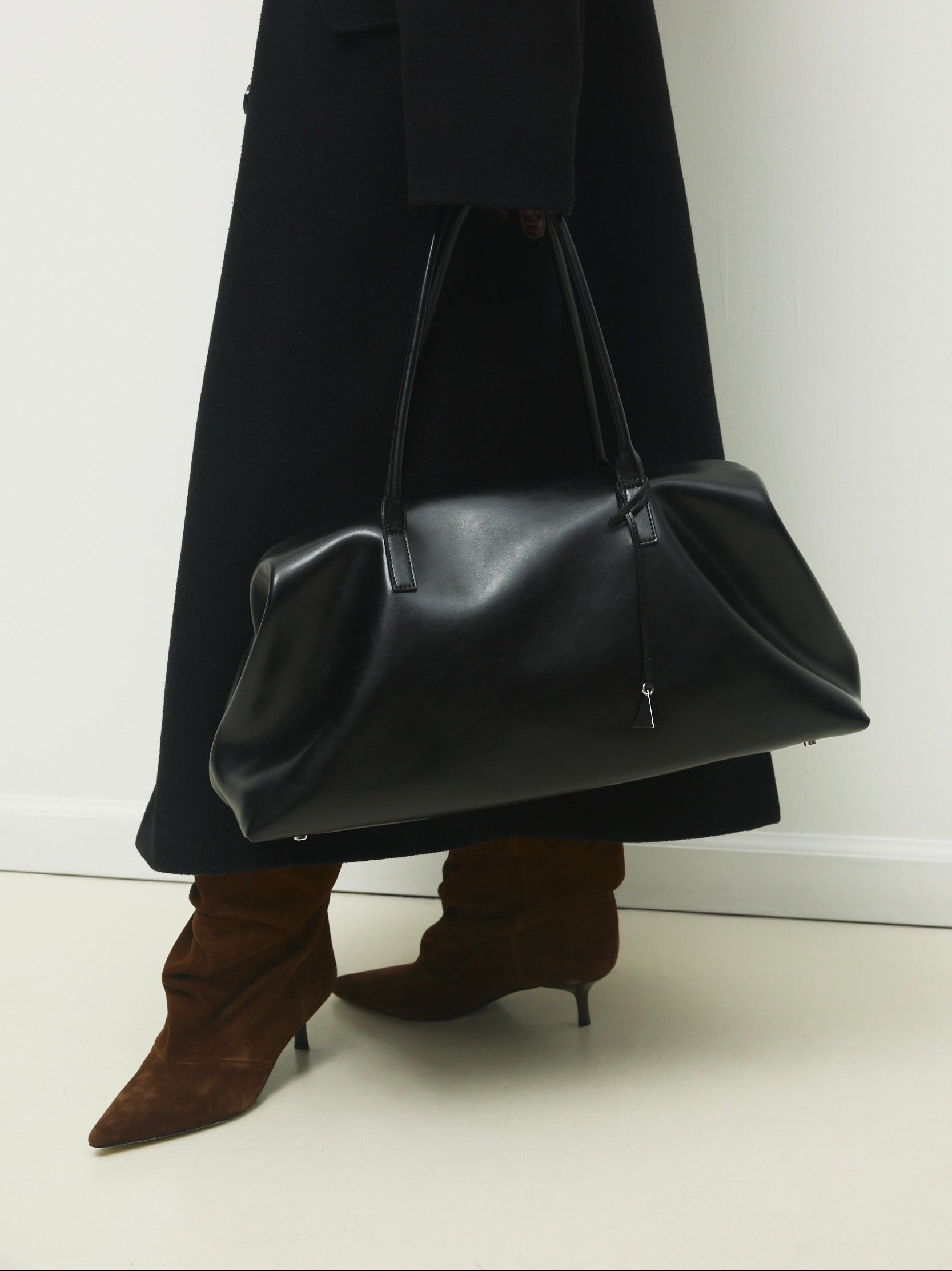 Person wearing a long black coat and brown boots holding a  Black Club Large Handbag against a white background
