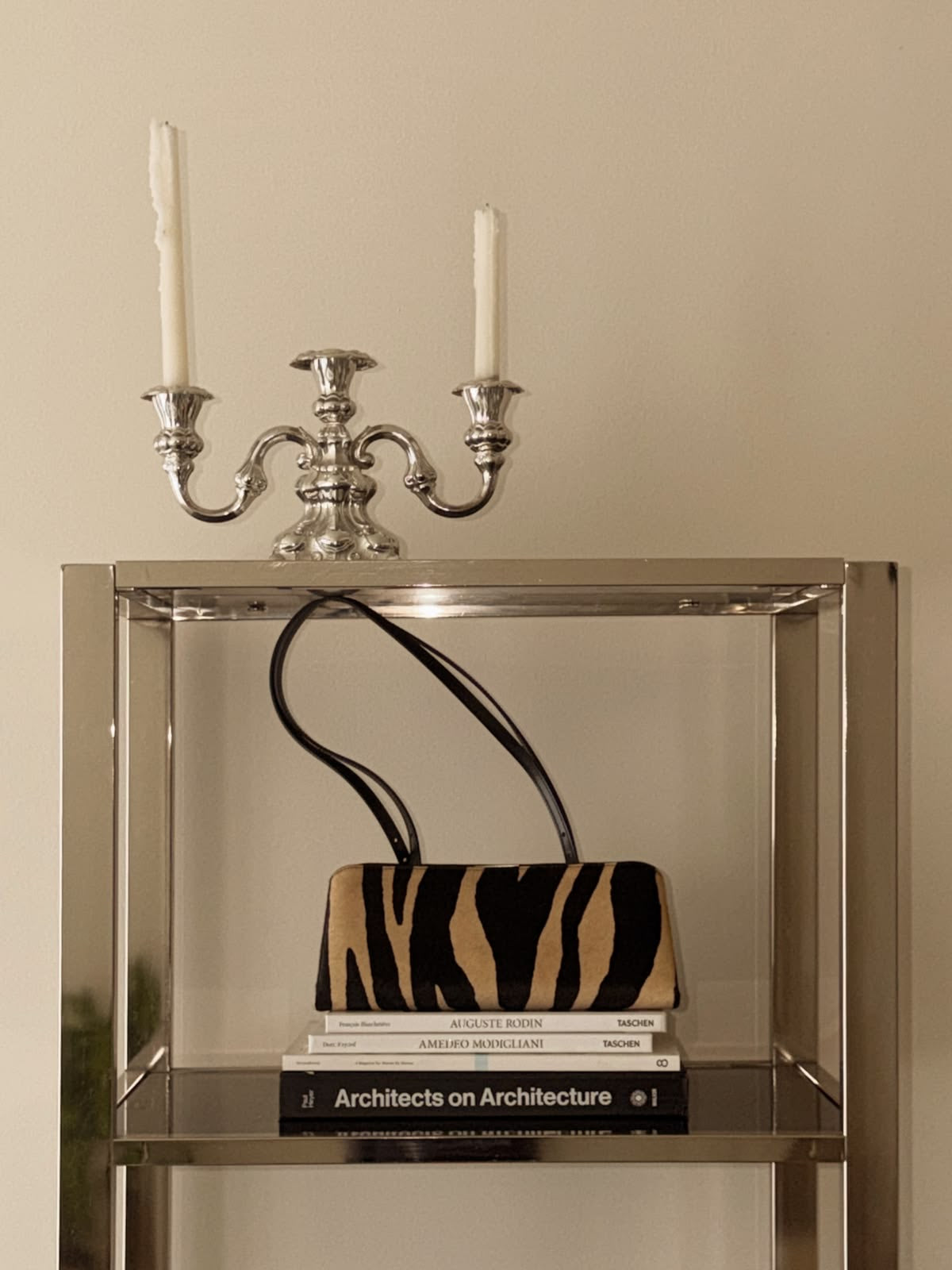 A structured handbag with a Zebra Calf Hair Café Mini Handbag, displayed on a glass shelf with books and a silver candelabra against a beige wall.