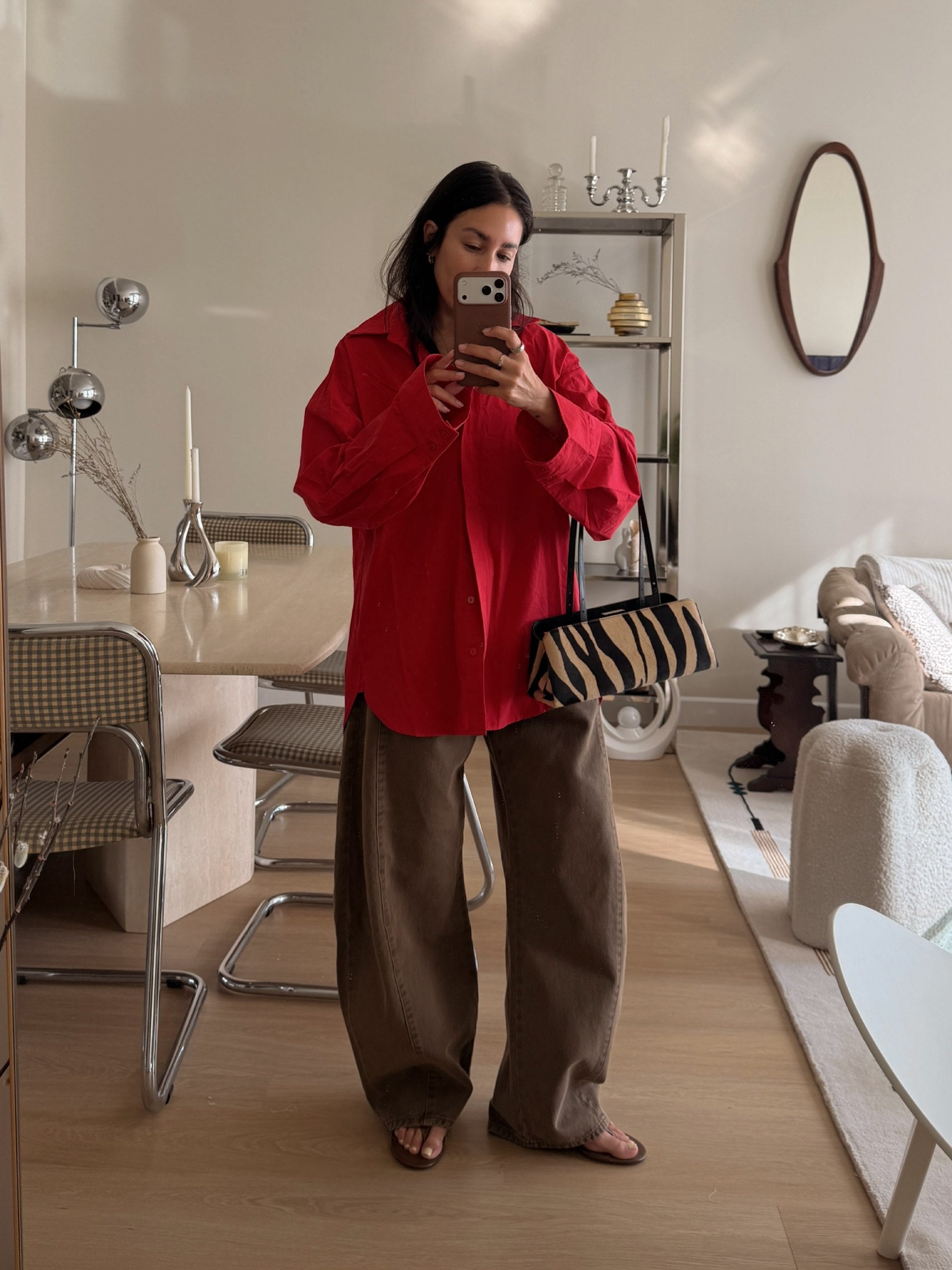A woman takes a mirror selfie in a bright, modern room, wearing a vibrant red button-up shirt, loose brown wide-leg trousers, and brown sandals, while holding a Zebra Calf Hair Café Mini Handbag.