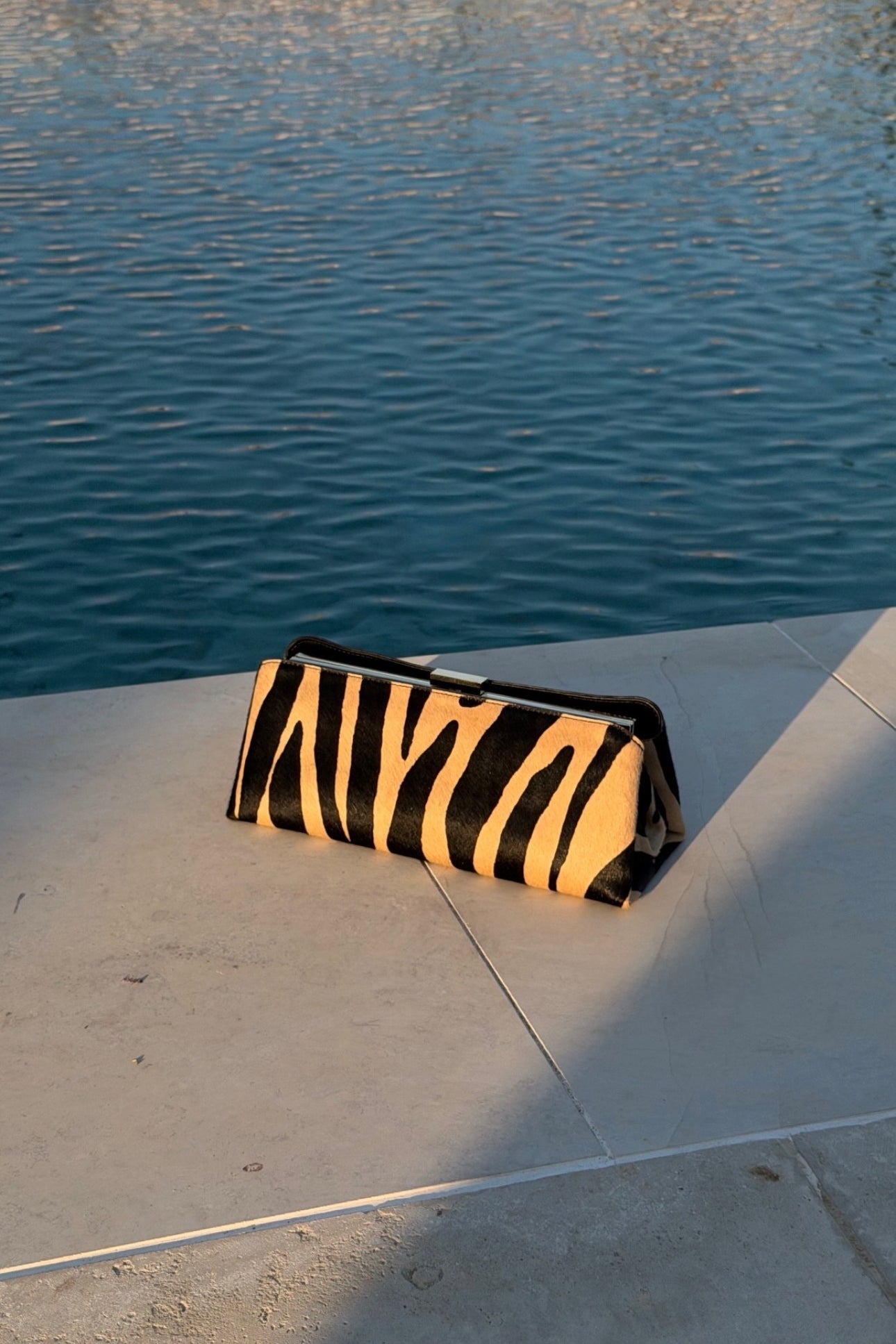 A Zebra Calf Hair Éclair Shoulder Handbag with black and tan stripes sits on a concrete ledge next to a swimming pool with blue water in the background.